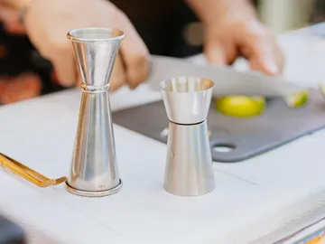 Zwei Edelstahl-Jigger stehen auf einem Tisch, während jemand schneidet.