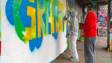 Zwei Personen sprühen bunte Graffiti-Wand an.