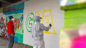 Zwei Personen sprühen Graffiti an Wand.