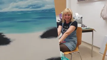 Woman smiling beside a beach painting indoors.