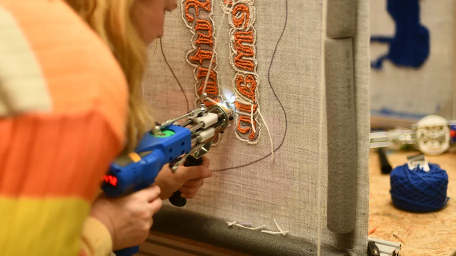 Person uses tufting gun on fabric frame.