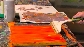 Person painting orange on canvas with spatula.