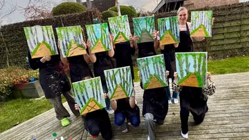Group holding paintings of forest path outdoors.