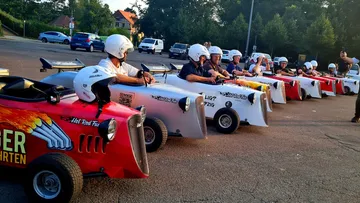 Group in race cars on asphalt, wearing helmets.