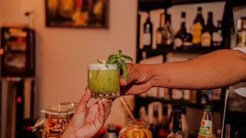 Zwei Hände tauschen einen grünen Cocktail aus.