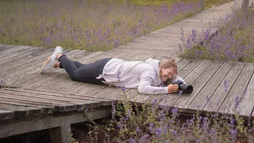 Person lying on wooden path photographing flowers.