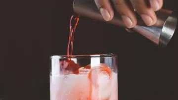Hand pouring red liquid into glass with ice.