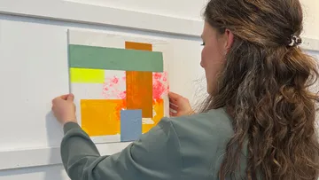 Woman hangs colorful abstract artwork on wall.