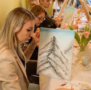 Women painting mountain landscapes at a table.