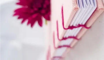 Rosafarbenes Buch mit rotem Faden, Blume daneben.