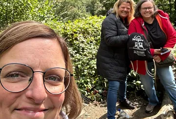 Drei Frauen im Freien, lächelnd, von Grün umgeben.
