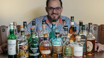 Mann lächelt, umgeben von verschiedenen Whiskeyflaschen.