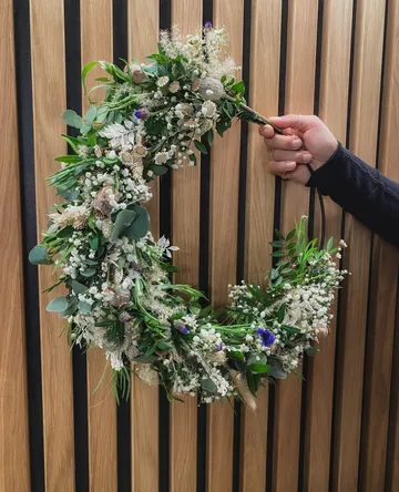 Hand hält Blumenkranz vor Holzwand.