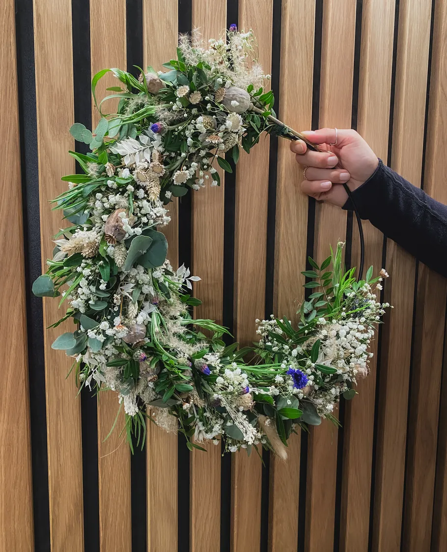 Hand hält Blumenkranz vor Holzwand.