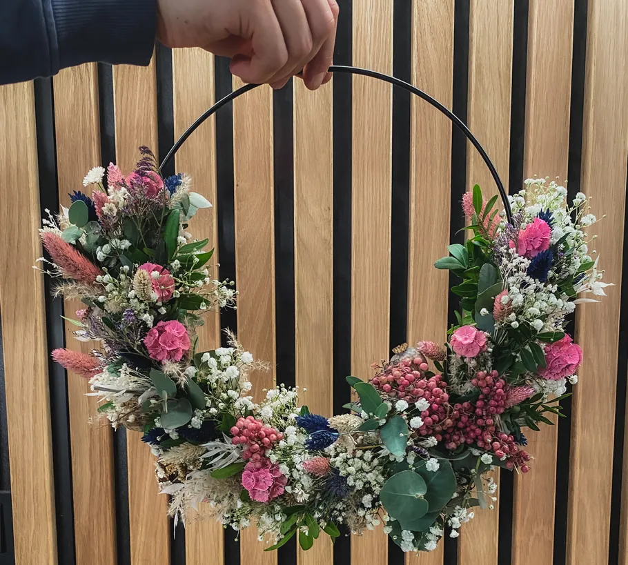 Hand hält Blumenkranz vor Holzwand.