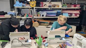 Three women sewing in fabric-filled room.