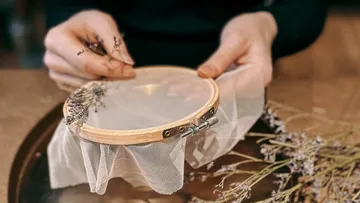 Hände sticken Lavendel auf Stoffrahmen.