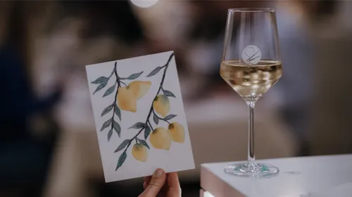 Hand holding painting of lemons, wine glass nearby.
