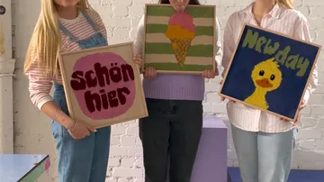 Drei Personen halten bunte gerahmte Kunstwerke drinnen.