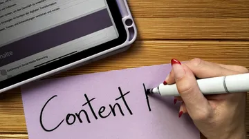 Hand schreibt auf Papier, Tablet daneben.