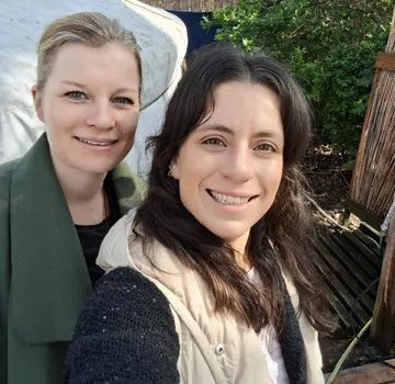 Zwei Frauen lächeln draußen bei Grünpflanzen.