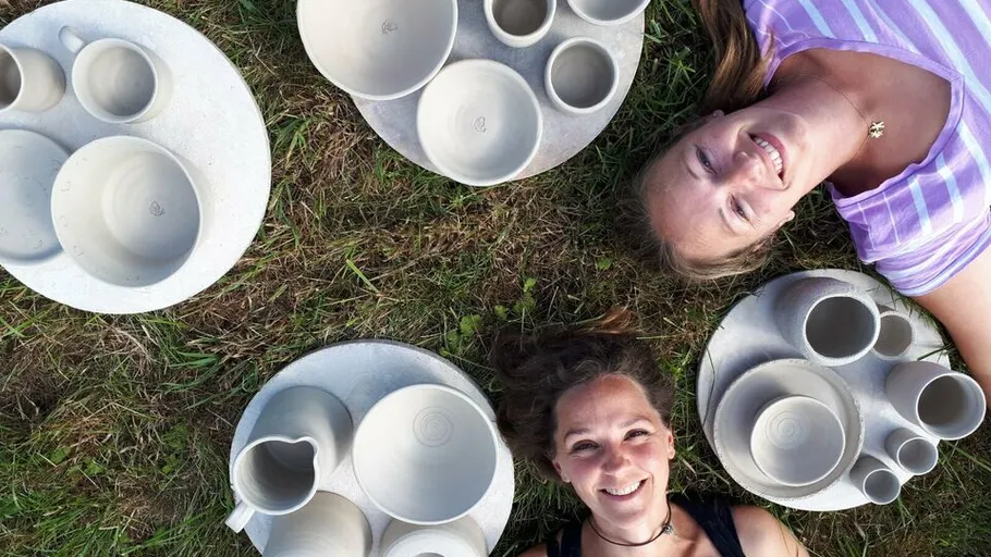 Zwei Frauen liegen auf Gras, umgeben von keramischen Geschirrteilen.