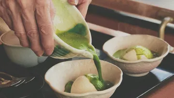 Hand pouring matcha sauce over ice cream bowls.