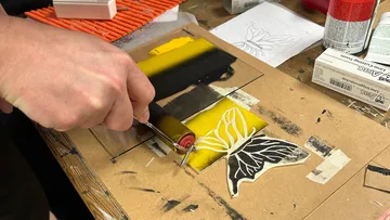Hand creating linocut butterfly print on yellow paper.