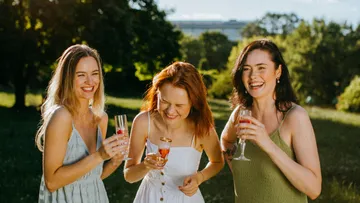 Three women laughing outdoors with drinks.