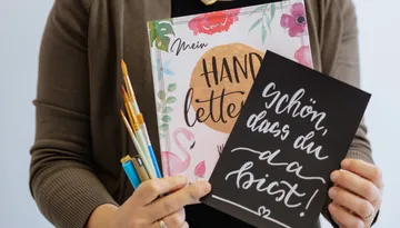Person hält Pinsel, Buch und Schild.