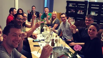 People raising glasses, sitting around table, indoors.