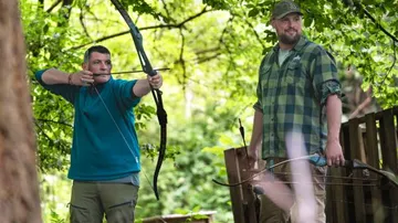 Two men practice archery in a forest.