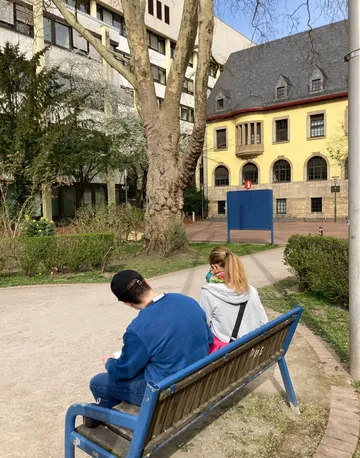 Personen sitzen auf Parkbank, Gebäude im Hintergrund.