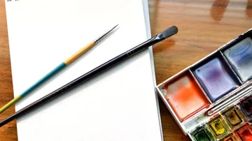 Paintbrushes, blank canvas, watercolor palette on table.