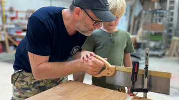Man teaching child to saw wood, workshop setting.
