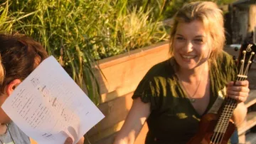 Woman with ukulele smiling in garden setting.
