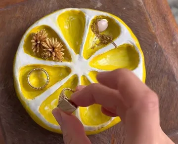 Hand hält Ring über zitronenförmige Schale mit Schmuck.