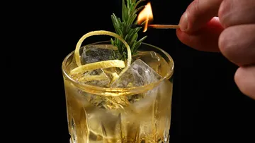 Cocktail mit Rosmarin-Garnitur, angezündetes Streichholz.
