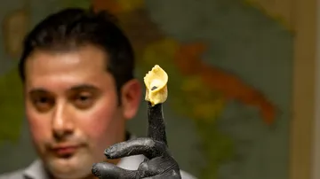 Man holding pasta on finger, blurry background.