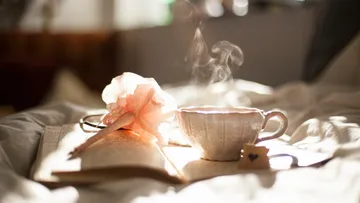 Dampfende Teetasse auf Bett mit Buch und Stift.