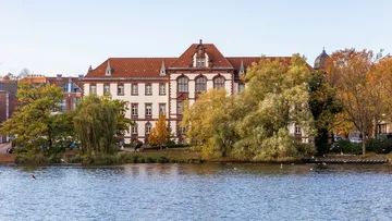 Historisches Gebäude am See, umgeben von Bäumen.