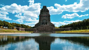 Denkmal spiegelt sich im Wasser, umgeben von Bäumen.