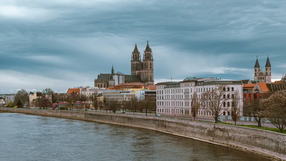 Riverside cityscape with cathedral under cloudy sky.