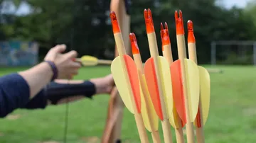 Pfeile im Vordergrund, Person zielt mit Bogen.