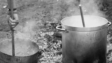 Hands stirring pots on outdoor fire.