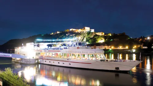 Illuminated boat on river with hills behind.