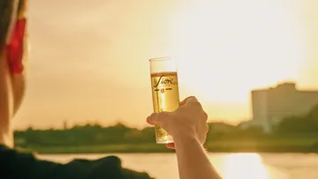 Person holding beer glass at sunset by river.