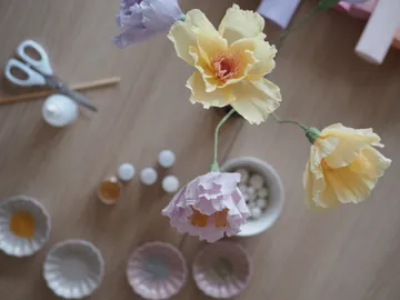 Bunte Papierblumen stehen auf einem Tisch mit Bastelmaterial.