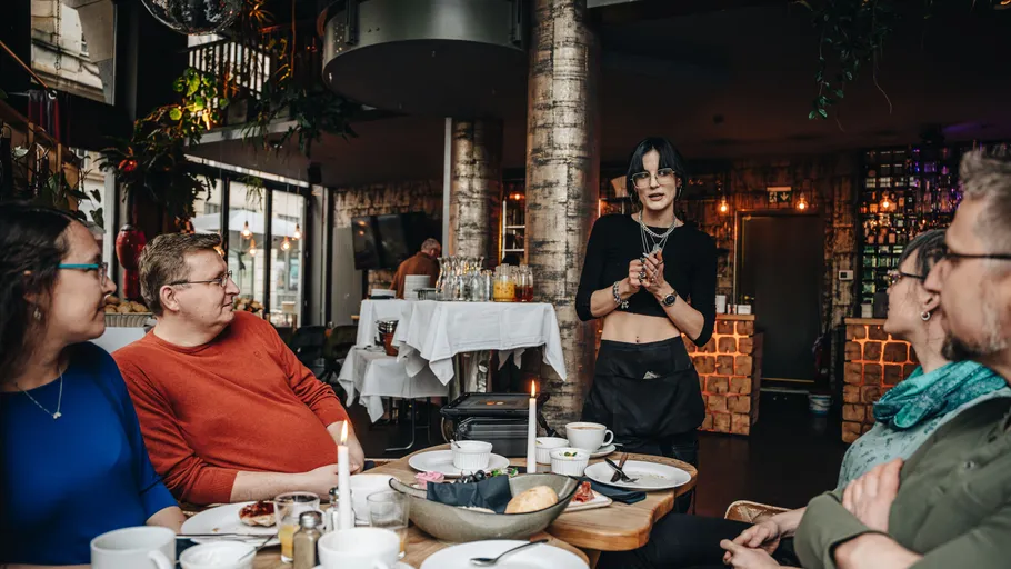 Woman standing, addressing seated diners in restaurant.