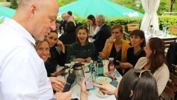 Mann mit Mikrofon spricht draußen mit sitzenden Frauen.